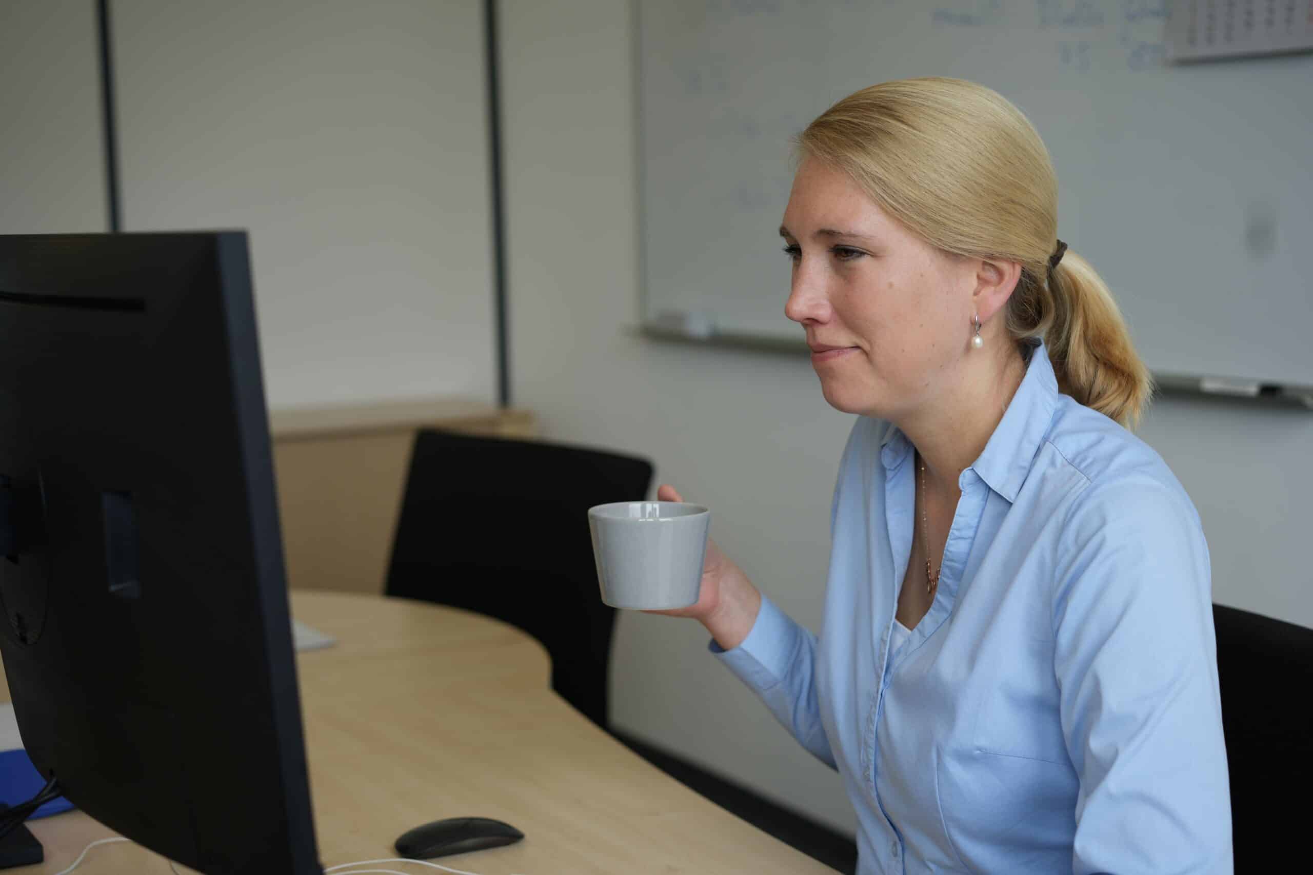Eine Frau schaut mit einem Kaffee in der Hand auf Ihren Bildschirm