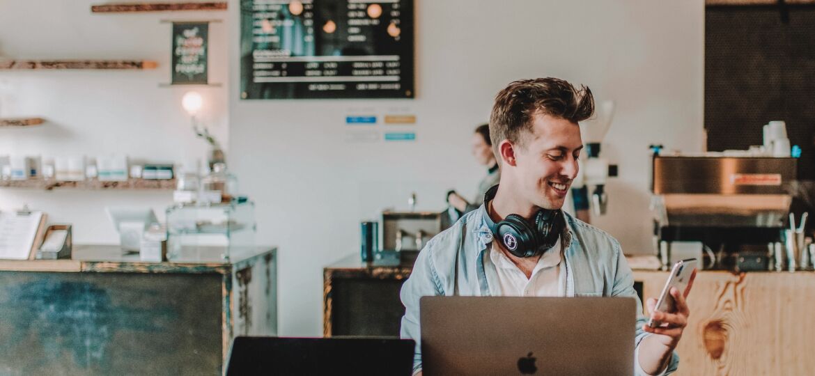 Ein Mann sitz in einem Café, vor ihm steht sein Laptop, er schaut lächelnd auf sein Handy