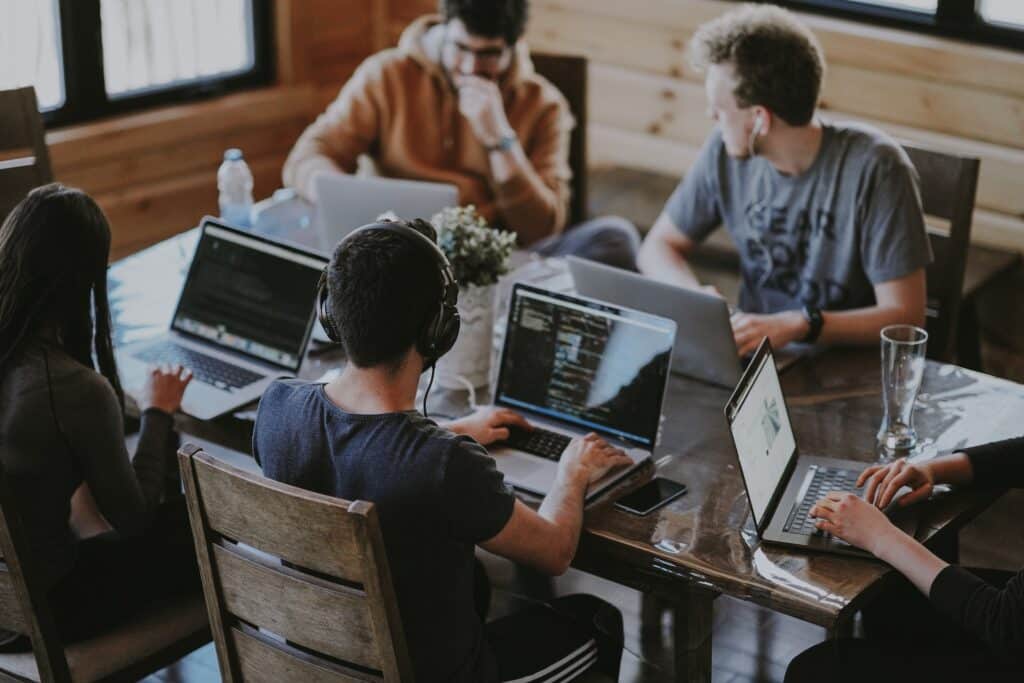 Produktivität steigern, Arbeitsbelastung senken Auf dem Foto sitzen fünf Menschen an einem Schreibtisch, alle haben einen Laptop vor sich und arbeiten