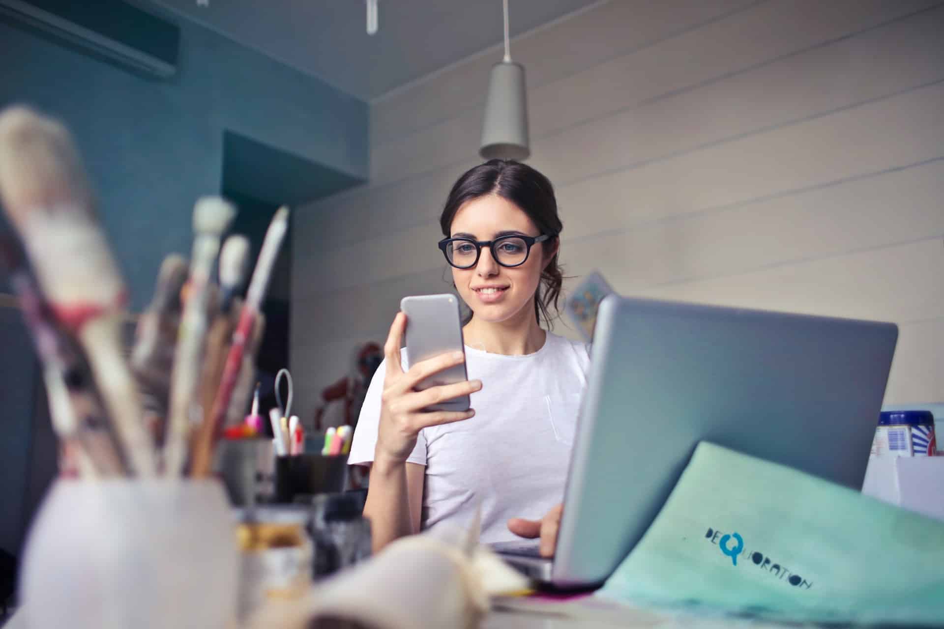 Eine Frau sitzt am Laptop und schaut auf ihr Handy