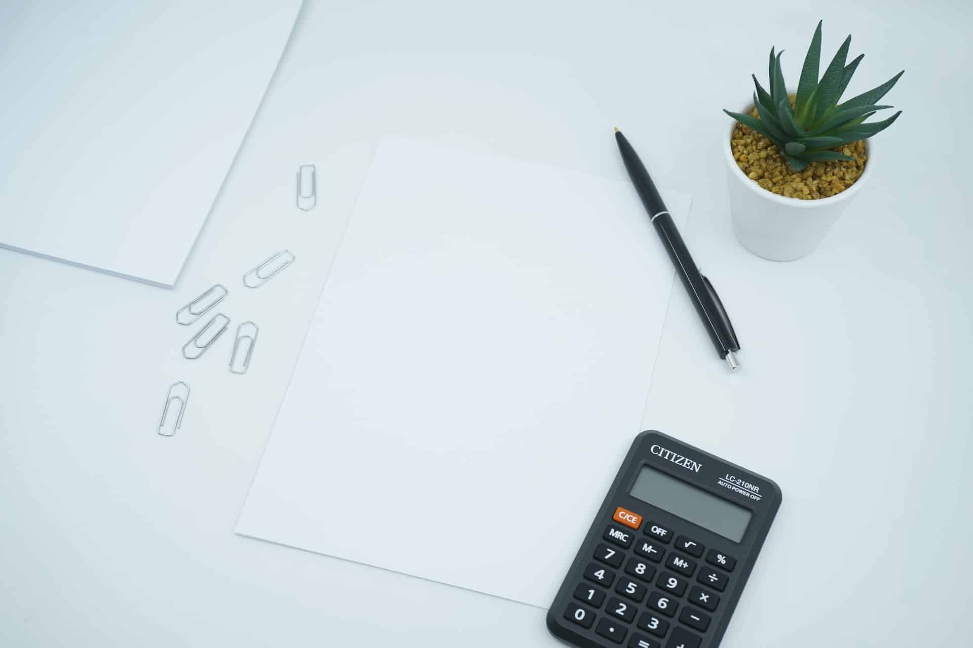 Auf einem Tisch liegen ein Blatt Papier, Büroklammern, ein Kulli und ein Taschenrechner