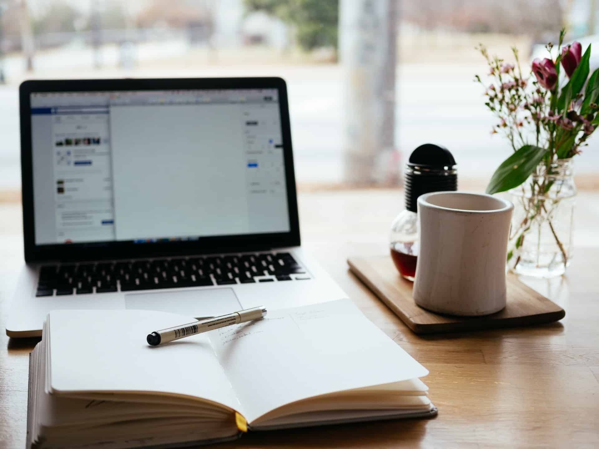 Ein Laptop, ein Notizbuch und eine Tasse stehen auf einem Schreibtisch