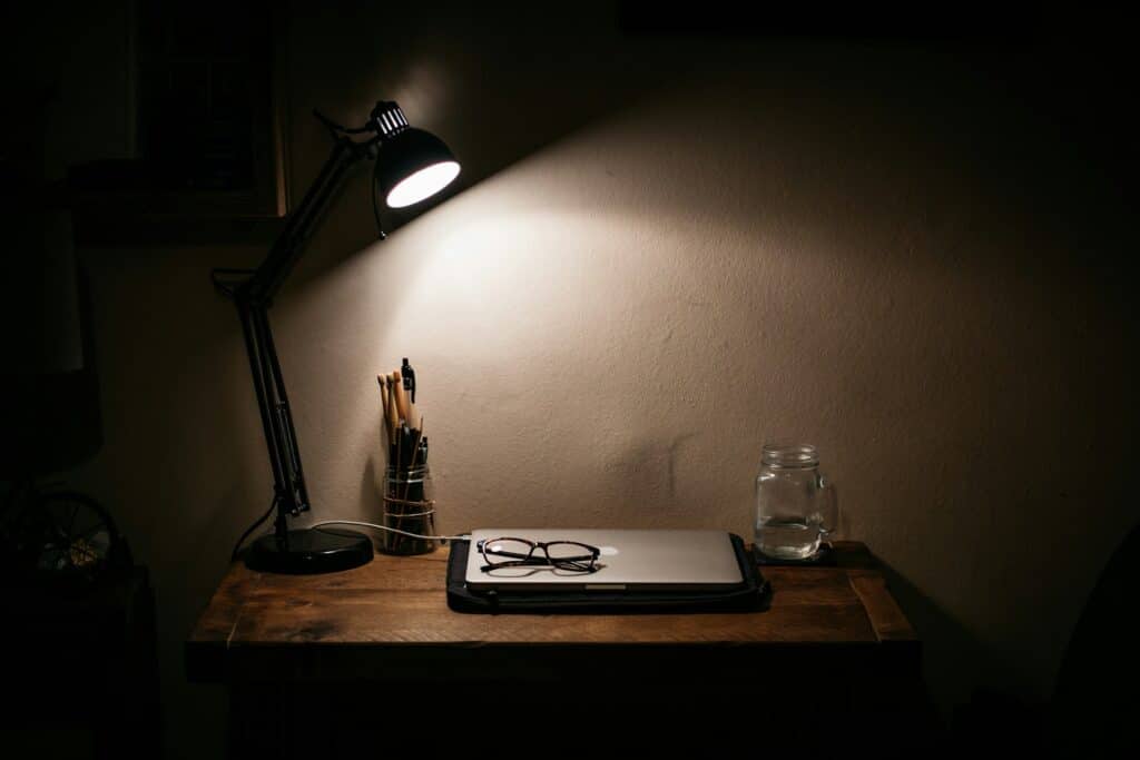 Ein Schreibtisch steht im Dunklen und wird nur mit einer Schreibtischleuchte beleuchtet. Auf dem Tisch steht ein zugeklappter Laptop, darauf eine Brille und daneben ein Glas Wasser