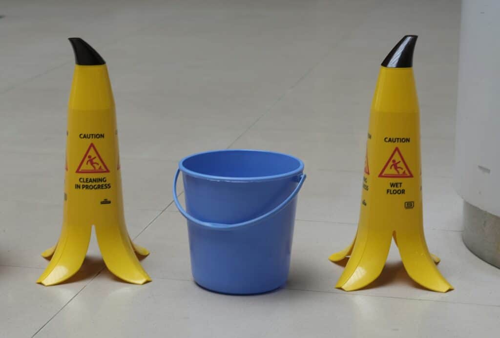 Zwei Schilder in Bananenform weisen auf nassen Boden hin. IN der Mitte steht ein blauer Putzeimer