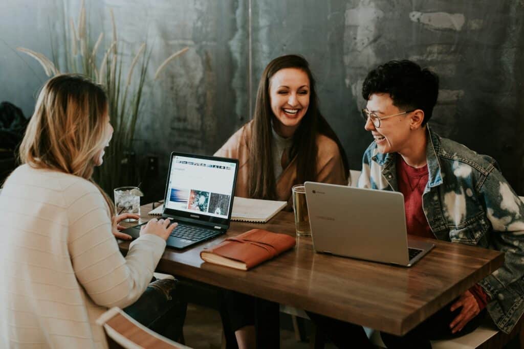 Drei Personen sitzen an einem Tisch vor ihren Laptops und arbeiten. Dabei reden sie und lachen.