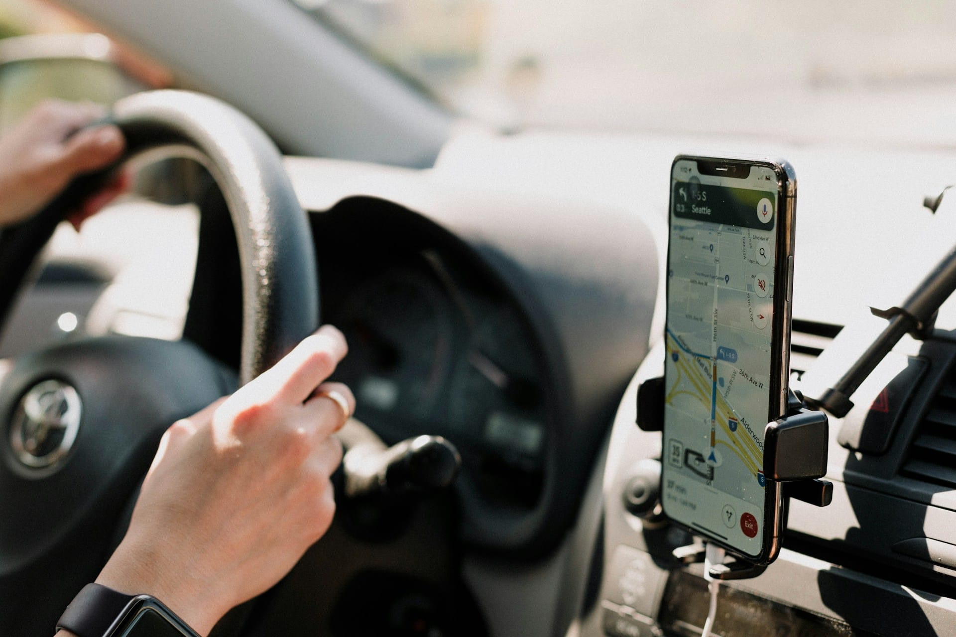 Eine Person sitz im Auto, im Ausschnitt zu sehen ist jedoch nur das Lenkrad und die Hände, die das Lenkrad umfassen. Neben dem Lenkrad ist einHandy befestigt, dass eine Wegbeschreibung abbildet