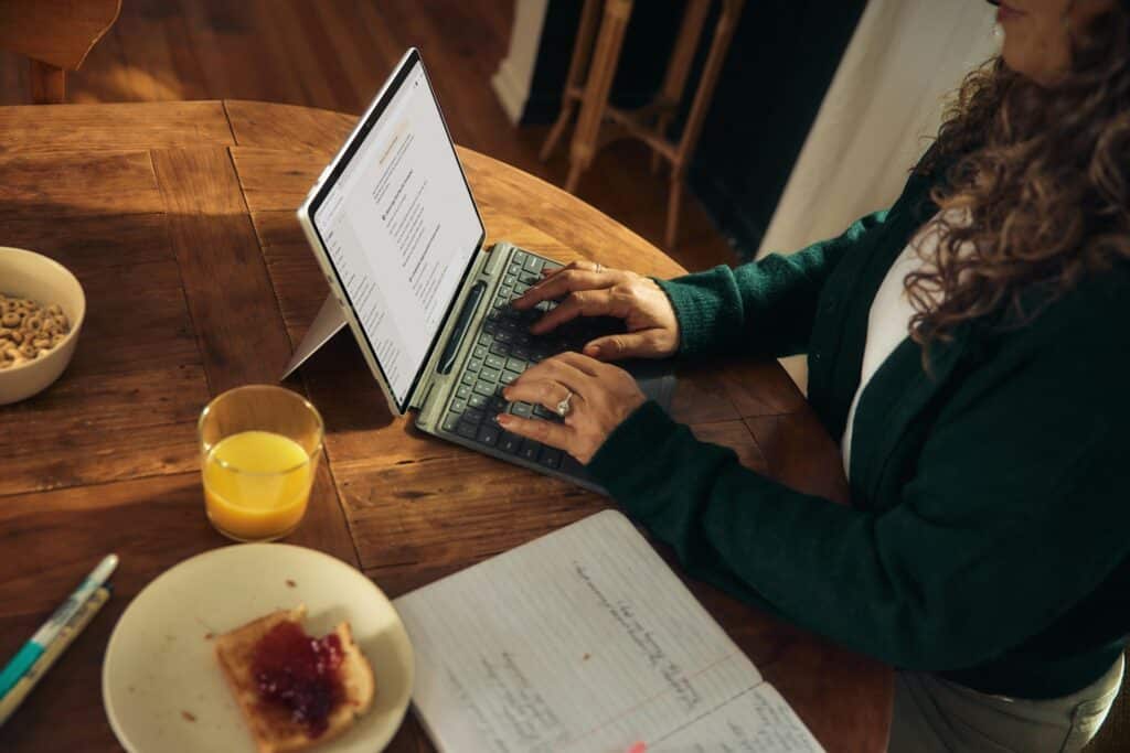 Eine Frau sitzt an einem Holztisch beim Frühstück und tippt auf einem Laptop